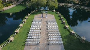 Aerial view of an elegant outdoor wedding ceremony setup with white chairs on green lawn beside a tranquil river.