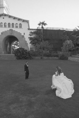 Black and white photo of bride in flowing gown and groom in tuxedo on lawn before elegant Spanish-style building.