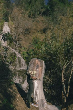 Bride and groom kissing under a veil near a waterfall surrounded by lush green trees on their wedding day.