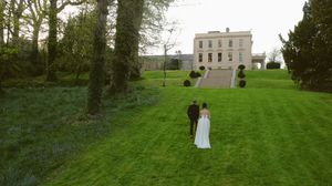 A scenic outdoor wedding ceremony setup with white chairs arranged on a lush green lawn surrounded by tall trees.
