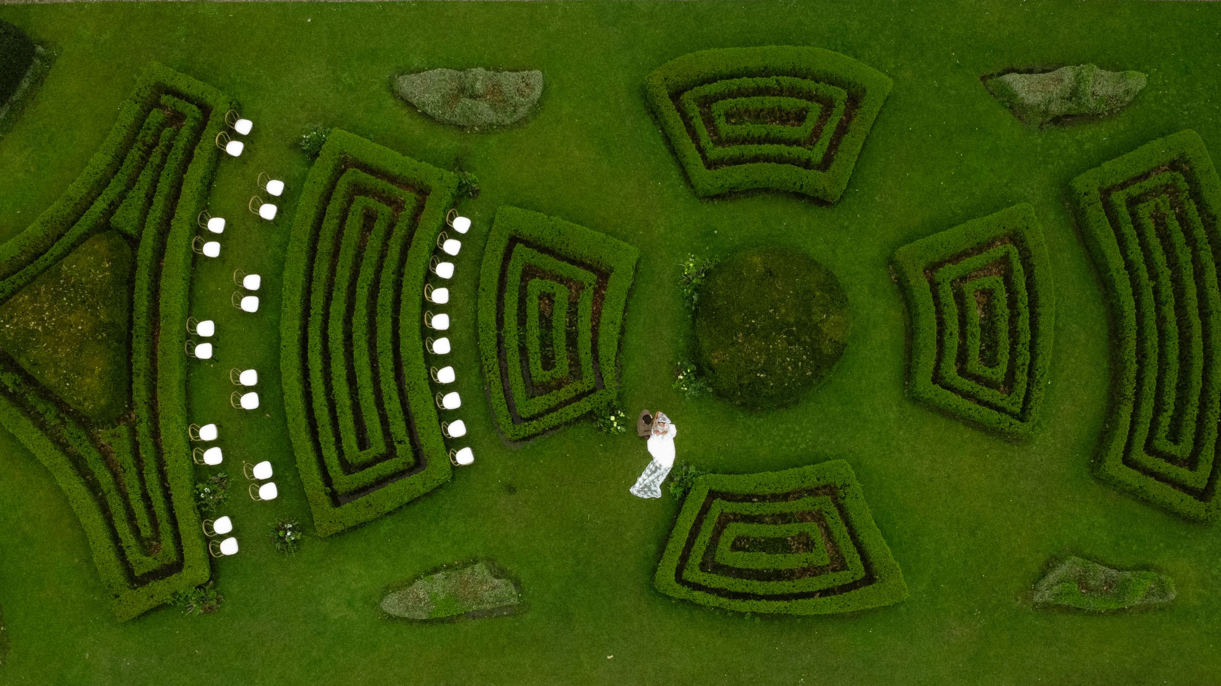 Aerial view of a symmetrical garden maze with geometric patterns and curved pathways on lush green grass.