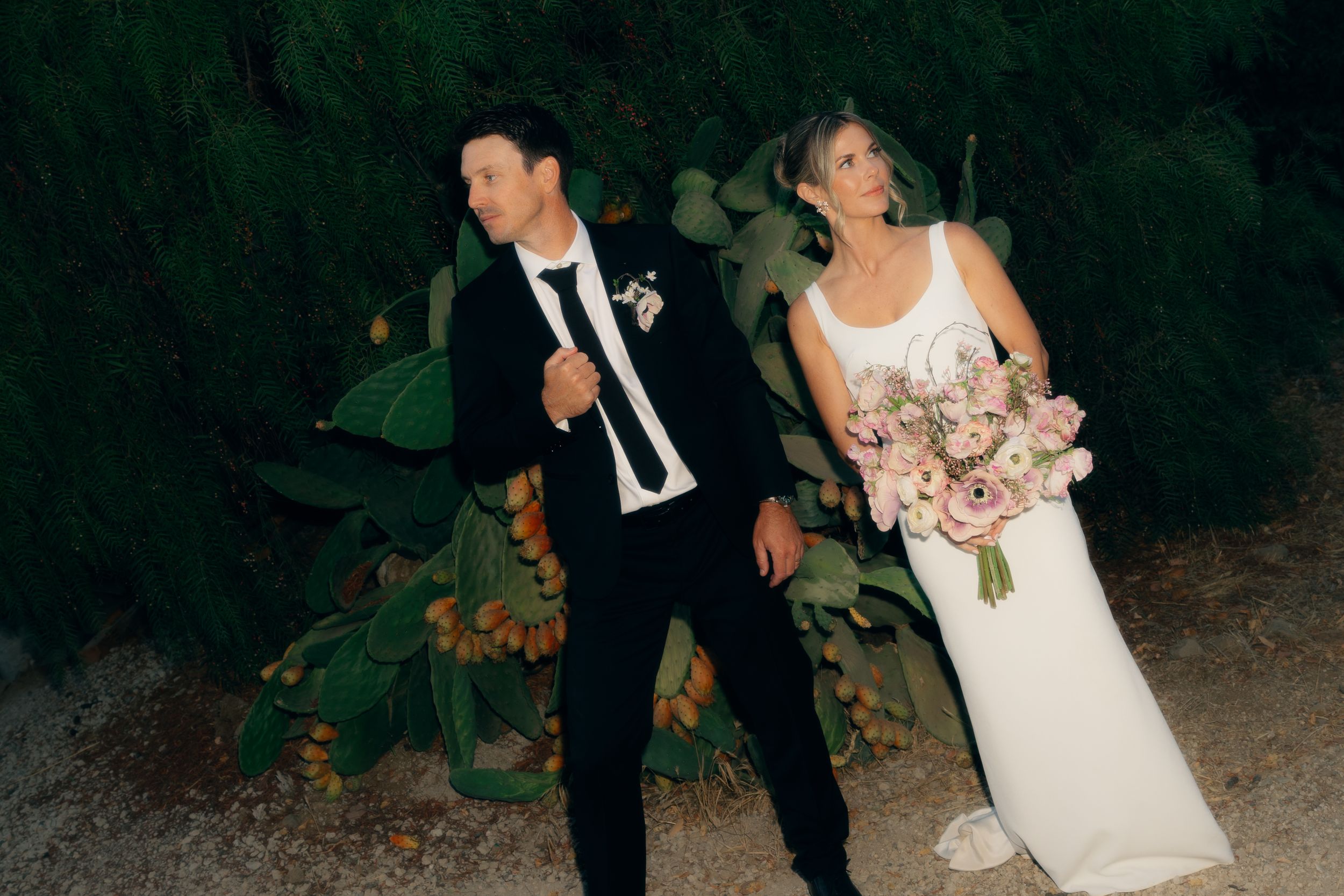Wedding party walks through a dark wooded area at night while holding bouquets and wearing formal attire.
