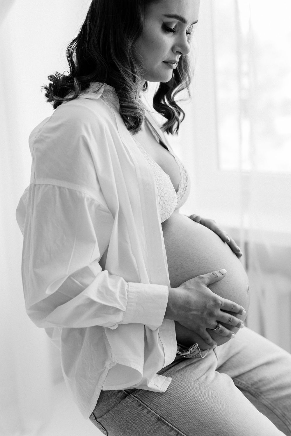 Black and white photo of pregnant woman in open white shirt tenderly holding her baby bump.