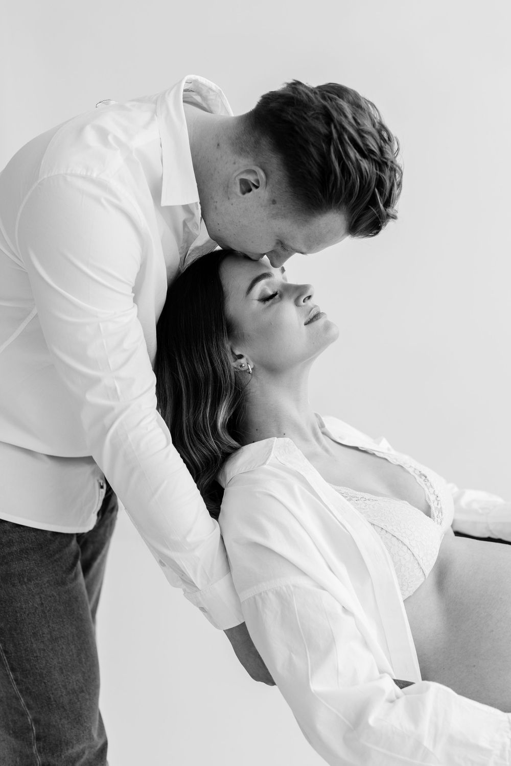 Black and white intimate photo of man leaning in to kiss pregnant woman in white studio setting.