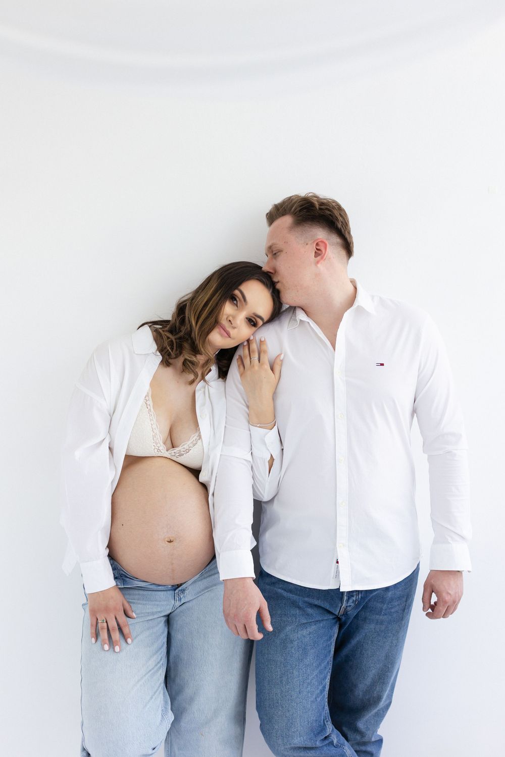 Couple in matching white shirts embrace, man kissing pregnant womans head against white wall.