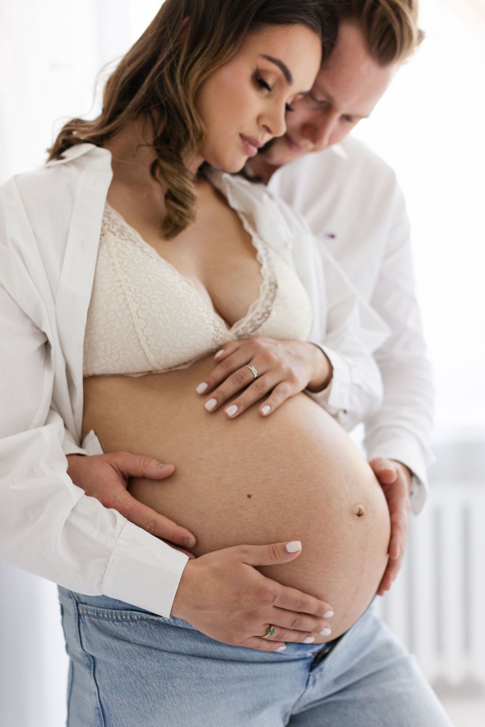 Man in white shirt tenderly cradles pregnant partners bare baby bump from behind near window.