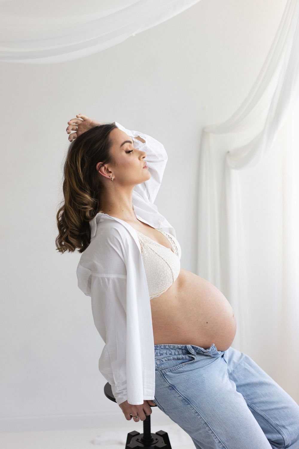 Pregnant woman in white shirt and jeans sits on stool, cradling baby bump in bright white studio.