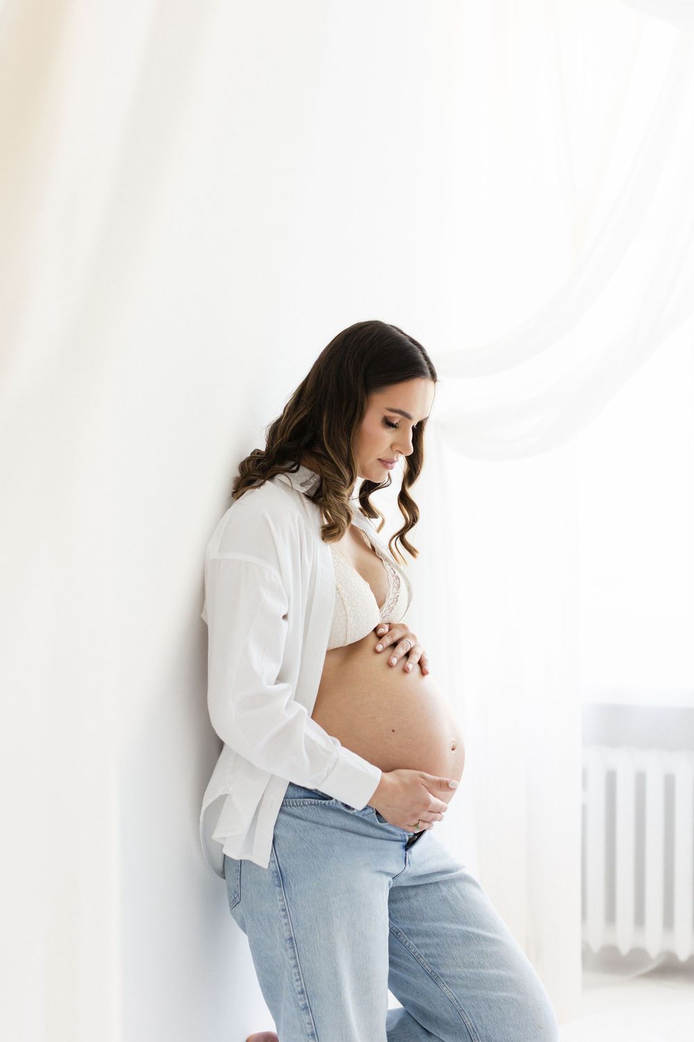 Pregnant woman in white shirt and jeans leans against white wall, gently touching her bare belly.