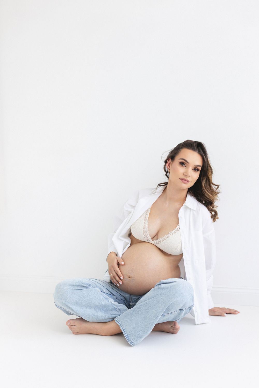 Pregnant woman sits cross-legged on white floor in open shirt and jeans, holding her baby bump.