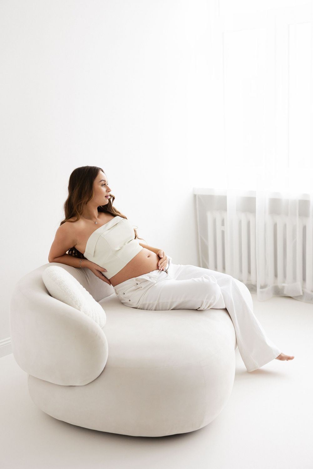 Smiling pregnant woman in white crop top and pants relaxes on round white chair in bright room.
