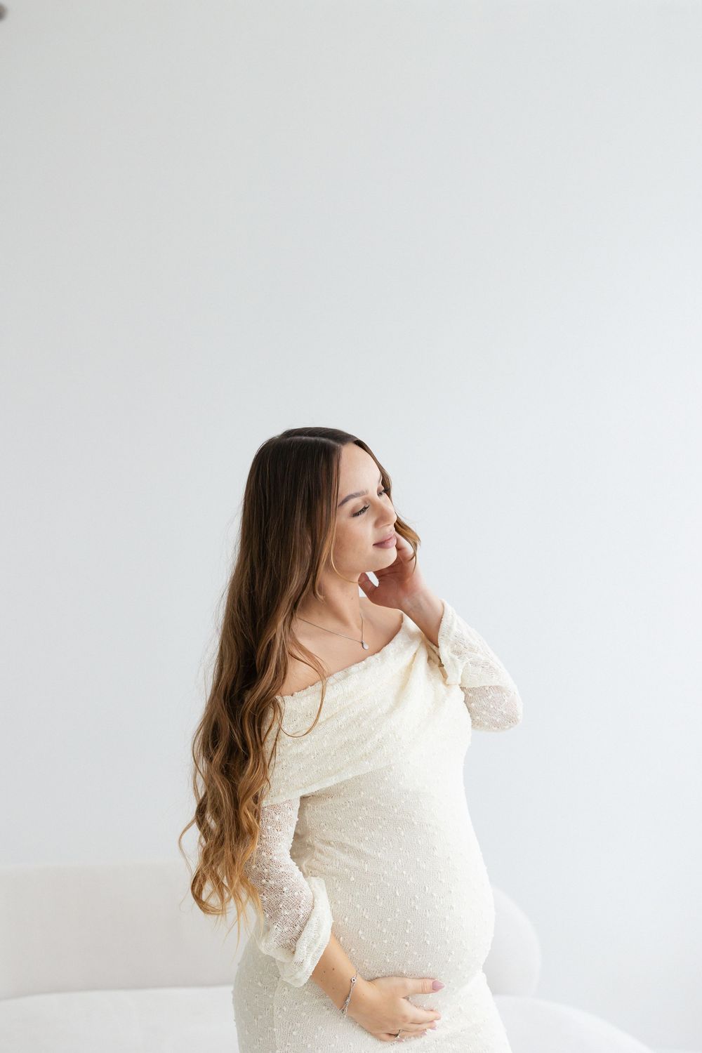 Pregnant woman in white lace dress touching her belly while looking away in bright studio.