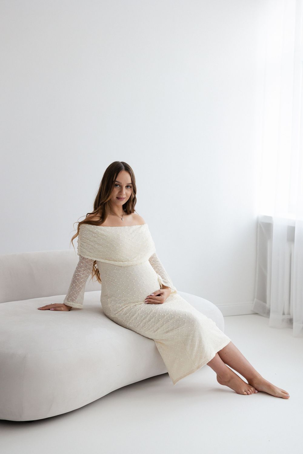 Smiling pregnant woman in off-shoulder white knit dress sitting on white sofa barefoot.
