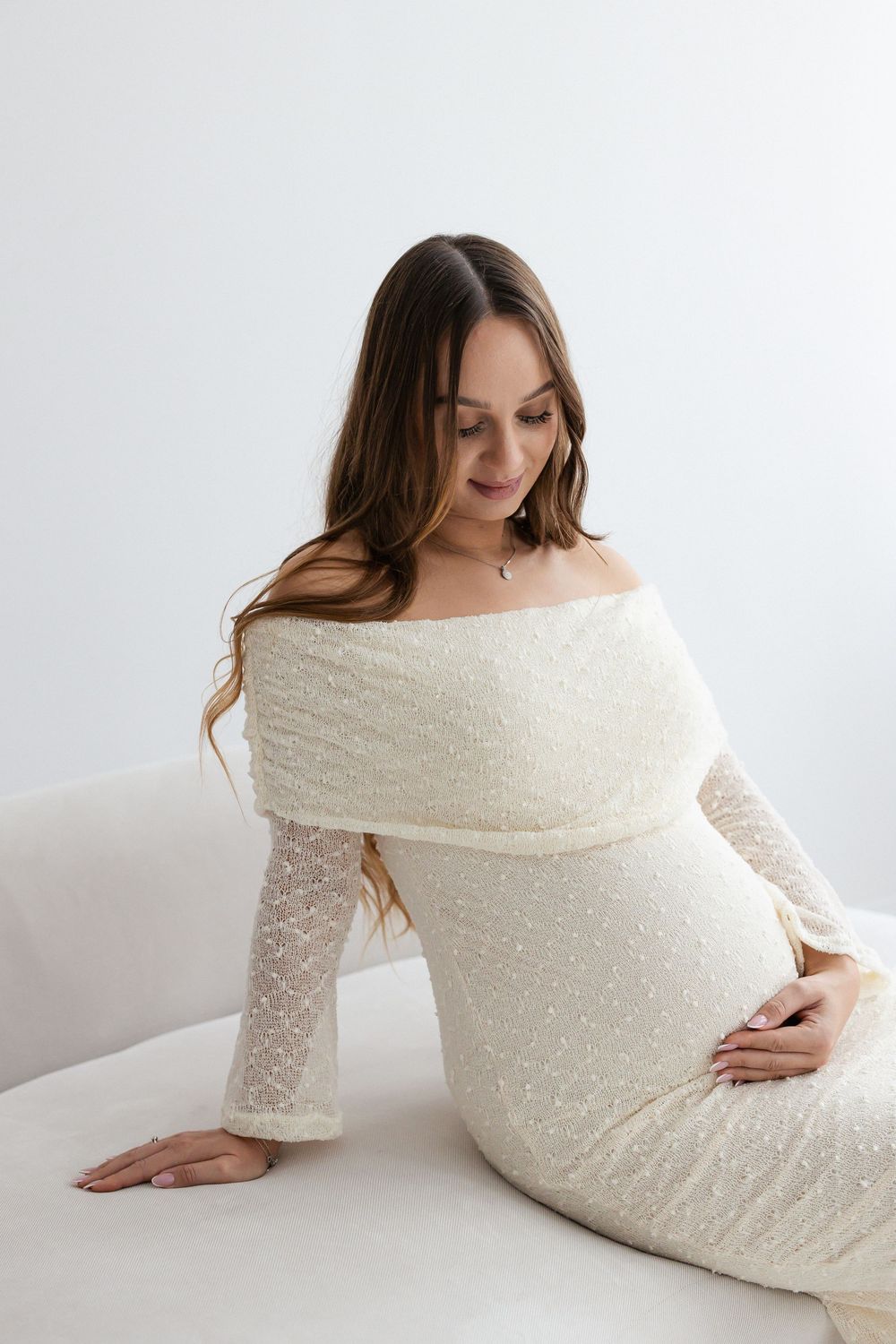 Pregnant woman in white off-shoulder dress looking down at belly while seated on sofa.