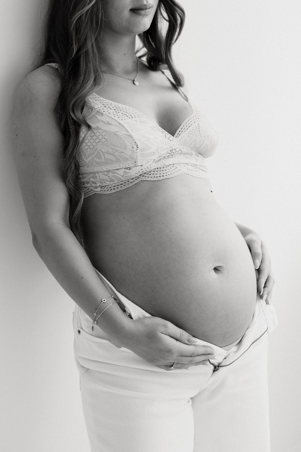 Black and white photo of pregnant woman in lace bra holding bare baby bump in white jeans.