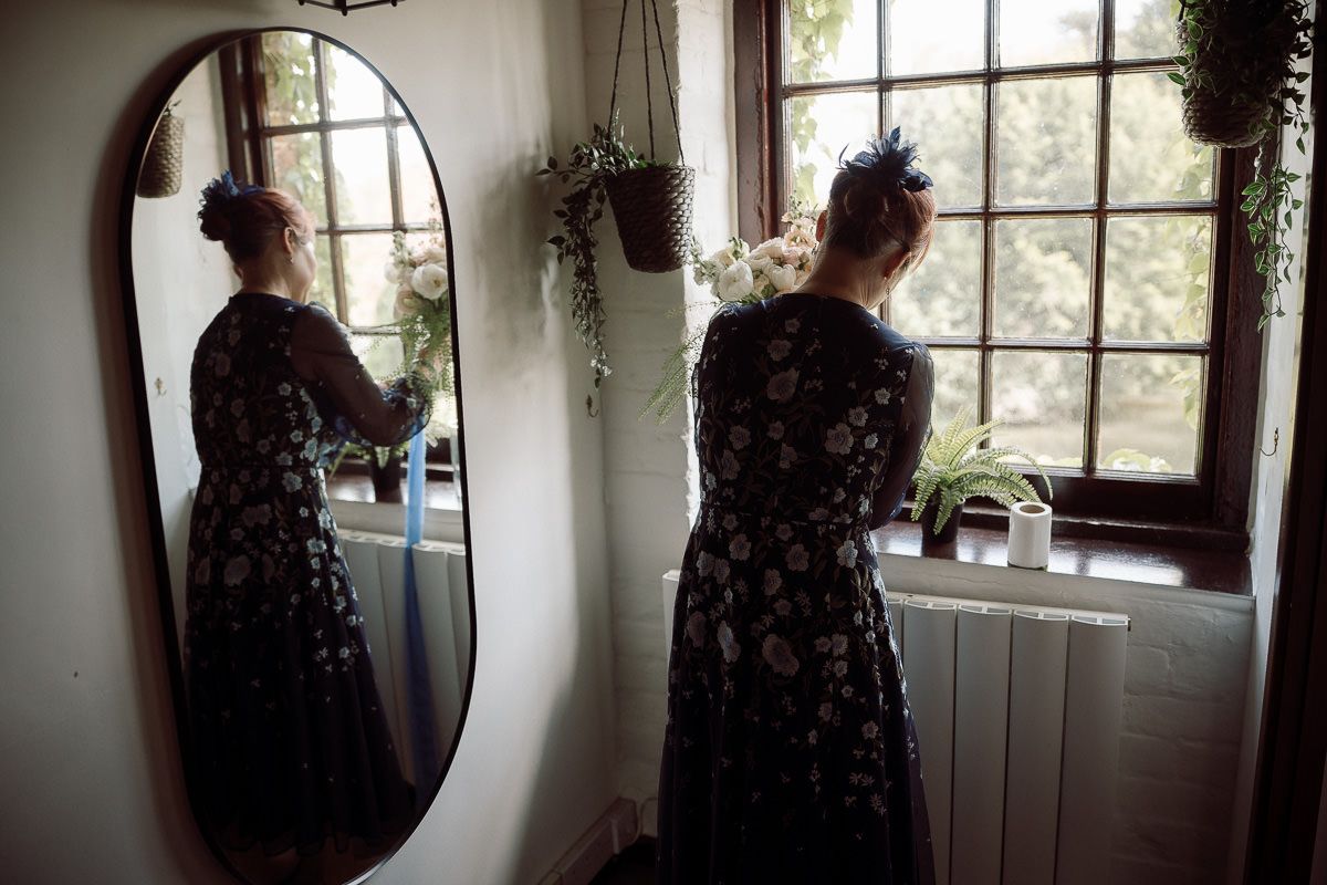 A person in a dark floral dress stands by a window in a rustic interior setting.