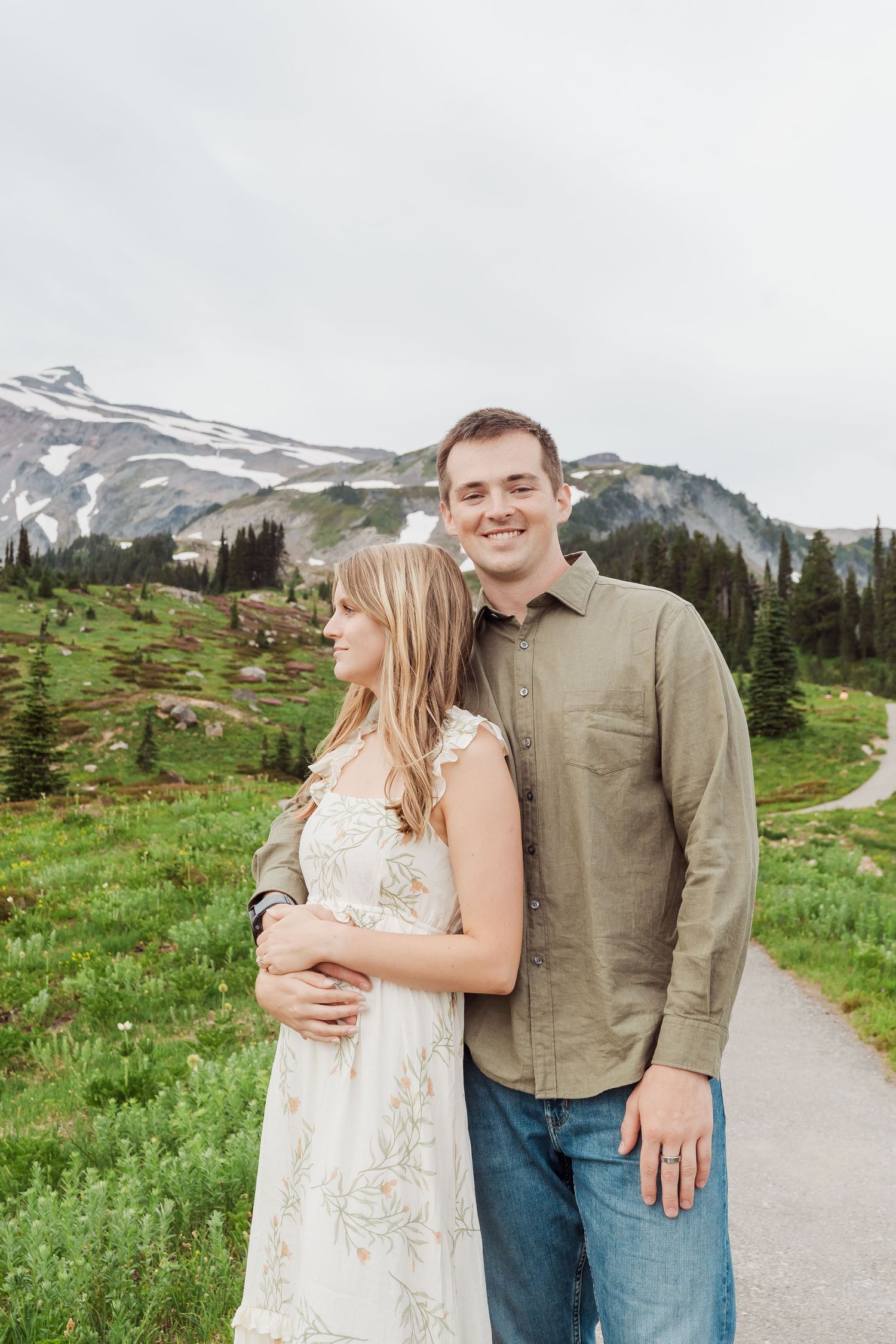 A couple embraces on a mountain trail surrounded by green meadows and snow-capped peaks in the background.