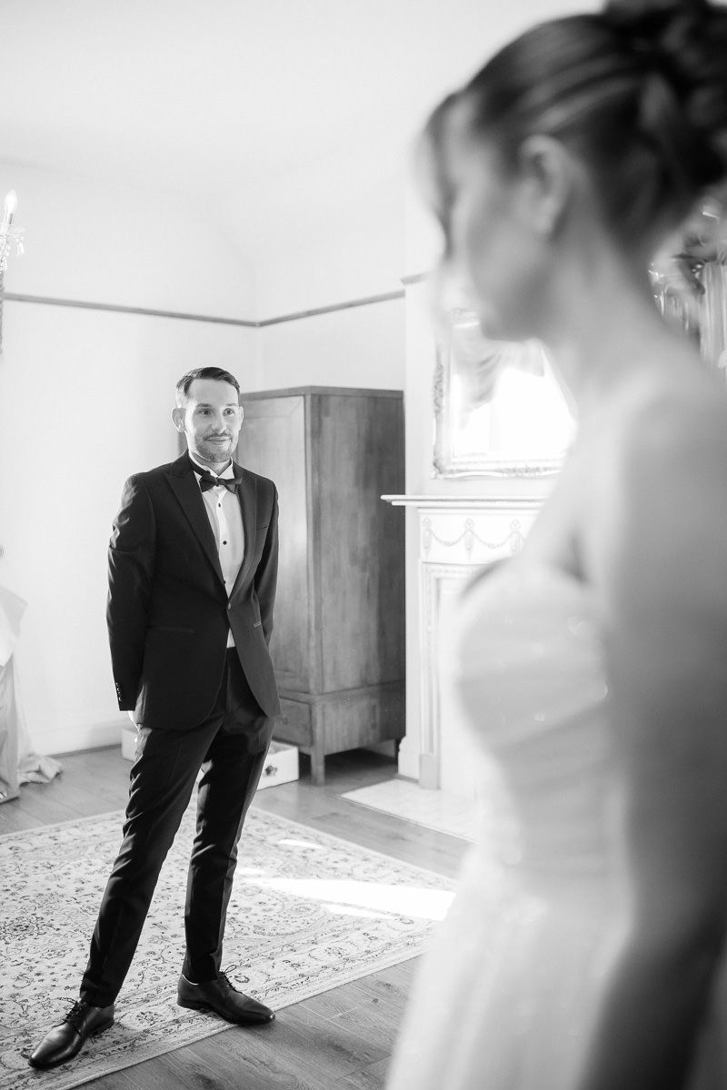 A black and white wedding photo series showing a groom in tuxedo standing in an elegant room with natural light.