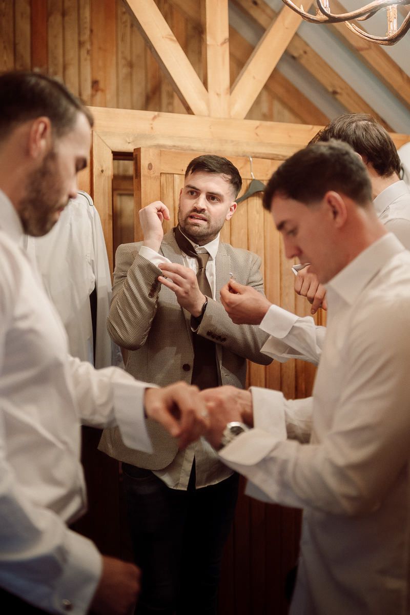 Groomsmen in white shirts helping each other get ready in a rustic wooden room.