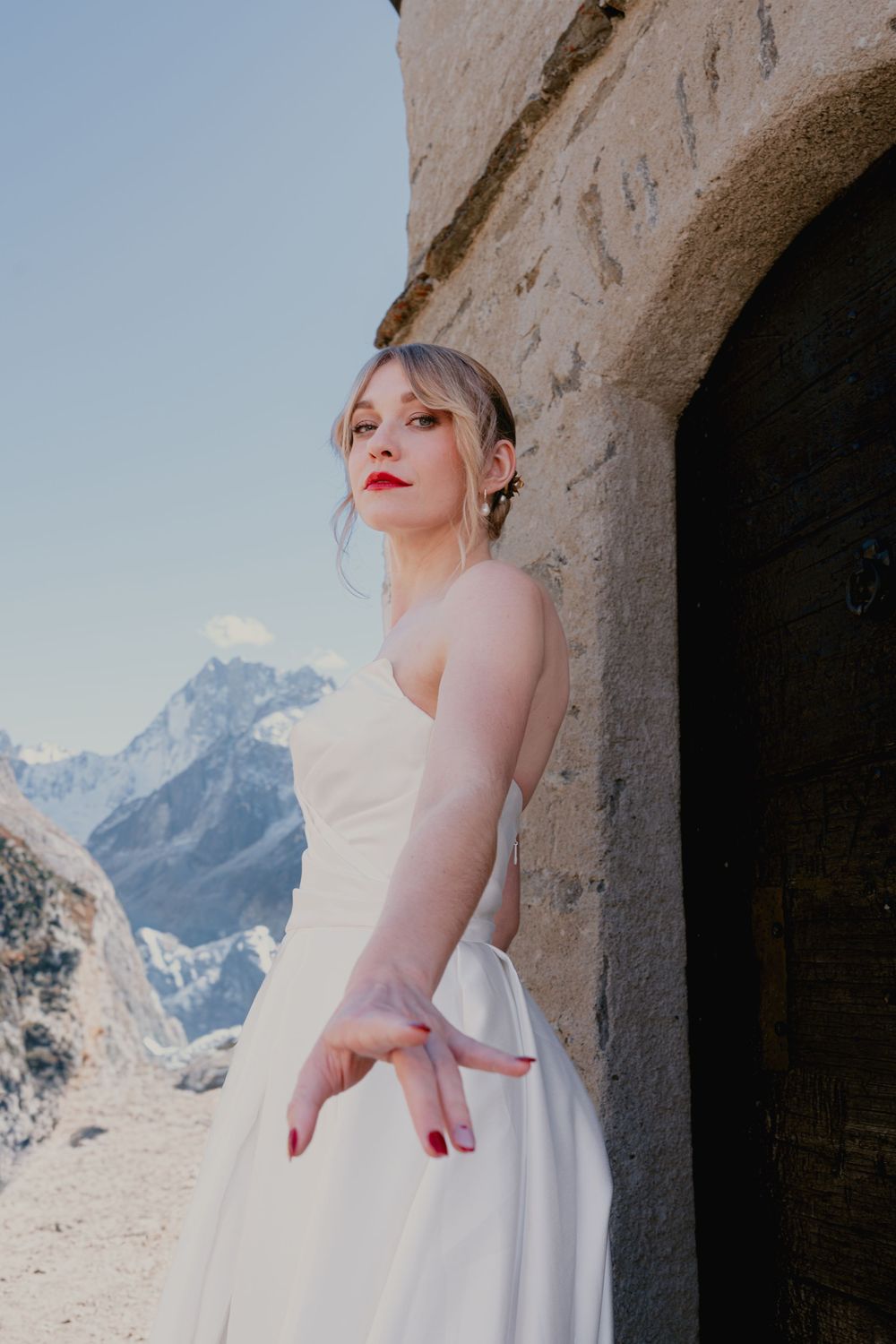 A woman in a white wedding dress poses against a stone archway with snow-capped mountains in the background.