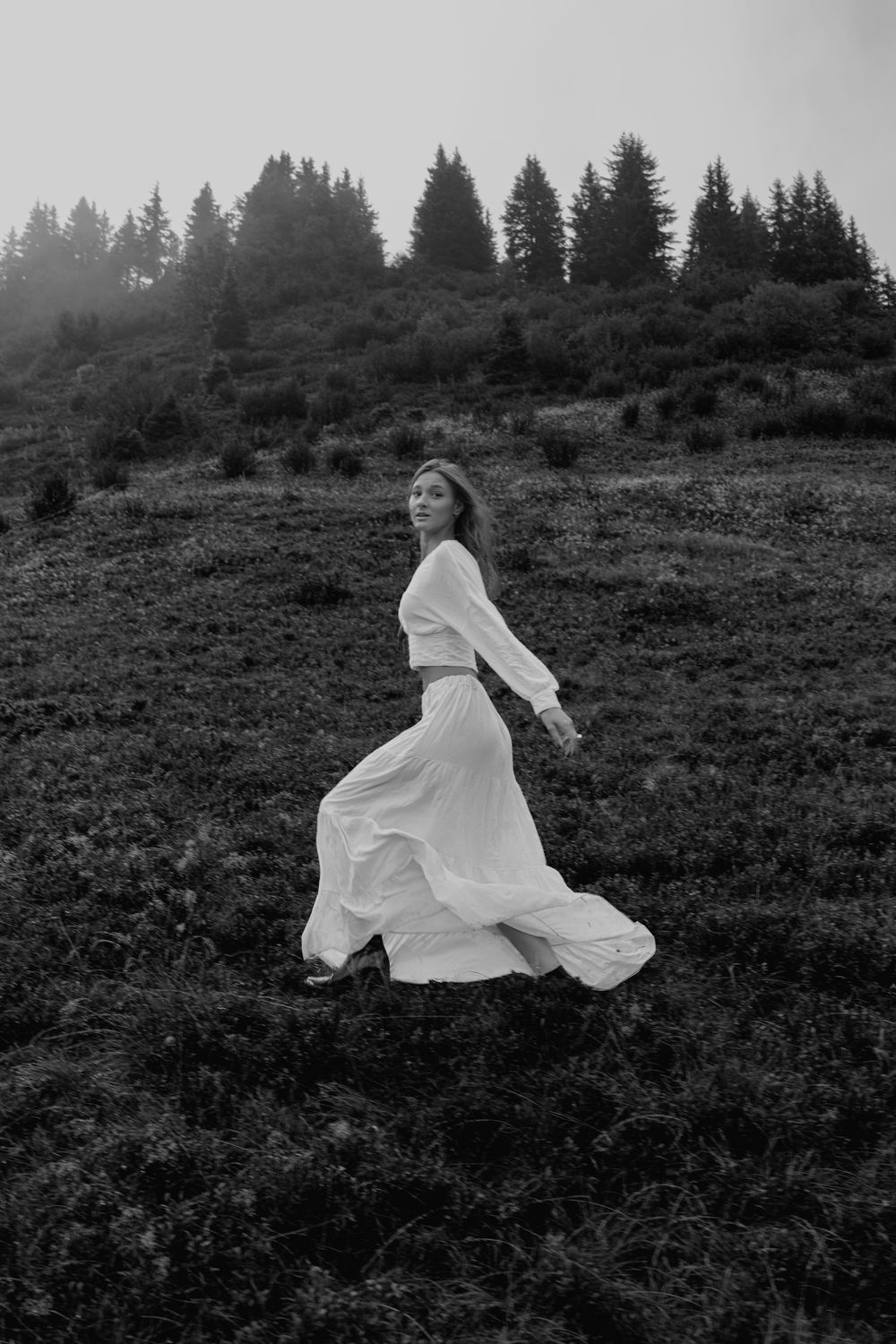 A figure in a flowing white dress moves gracefully across a misty hillside with evergreen trees in the background.