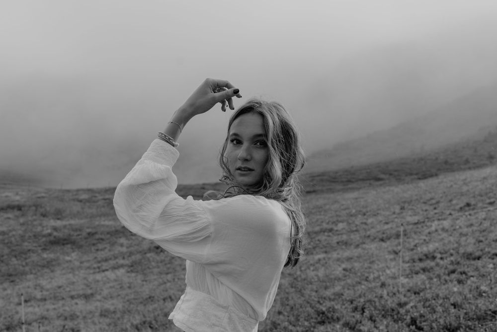 A person in a white dress stands on a misty mountainside while looking into the distance.