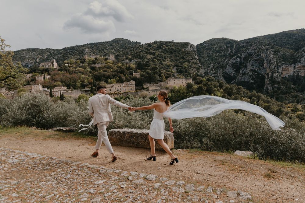 A couple walks along a mountain path while a flowing white dress catches the wind against a scenic landscape backdrop.