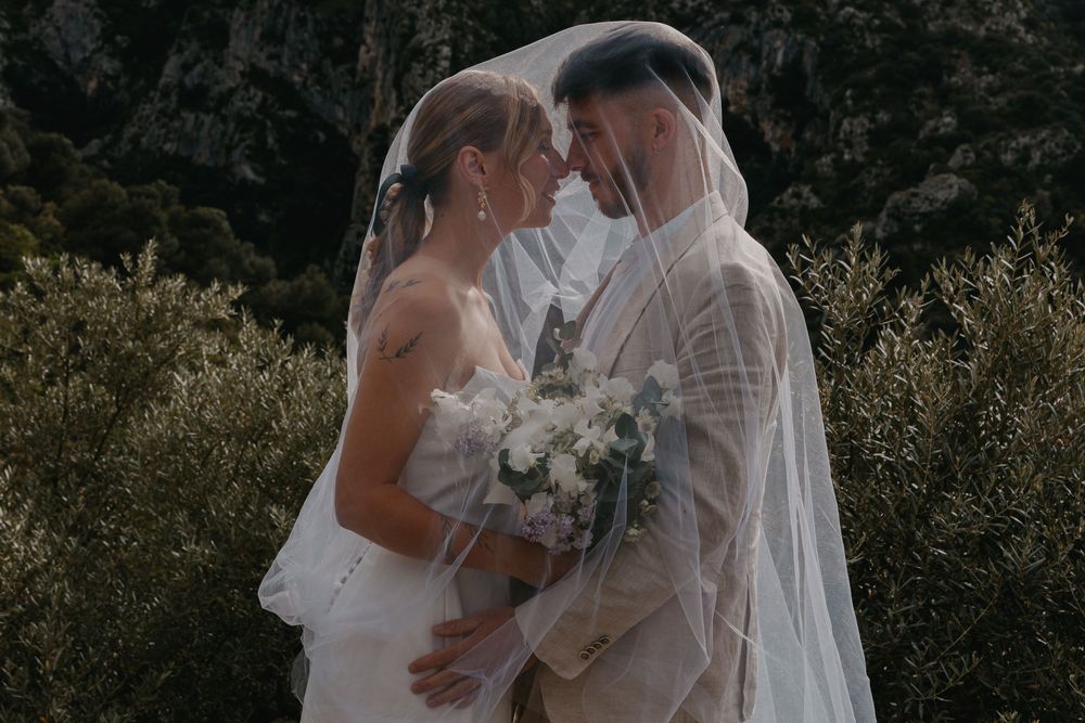 A couple in wedding attire shares an intimate moment outdoors surrounded by lush greenery and natural light.