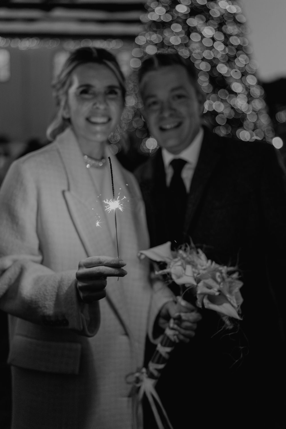 A black and white wedding photo with sparklers and twinkly Christmas lights creating a magical holiday atmosphere.