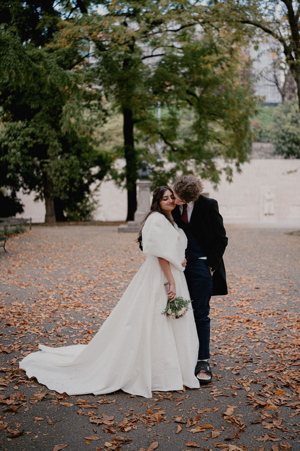 A couple shares a romantic moment on a path covered in fallen autumn leaves while wearing wedding attire.