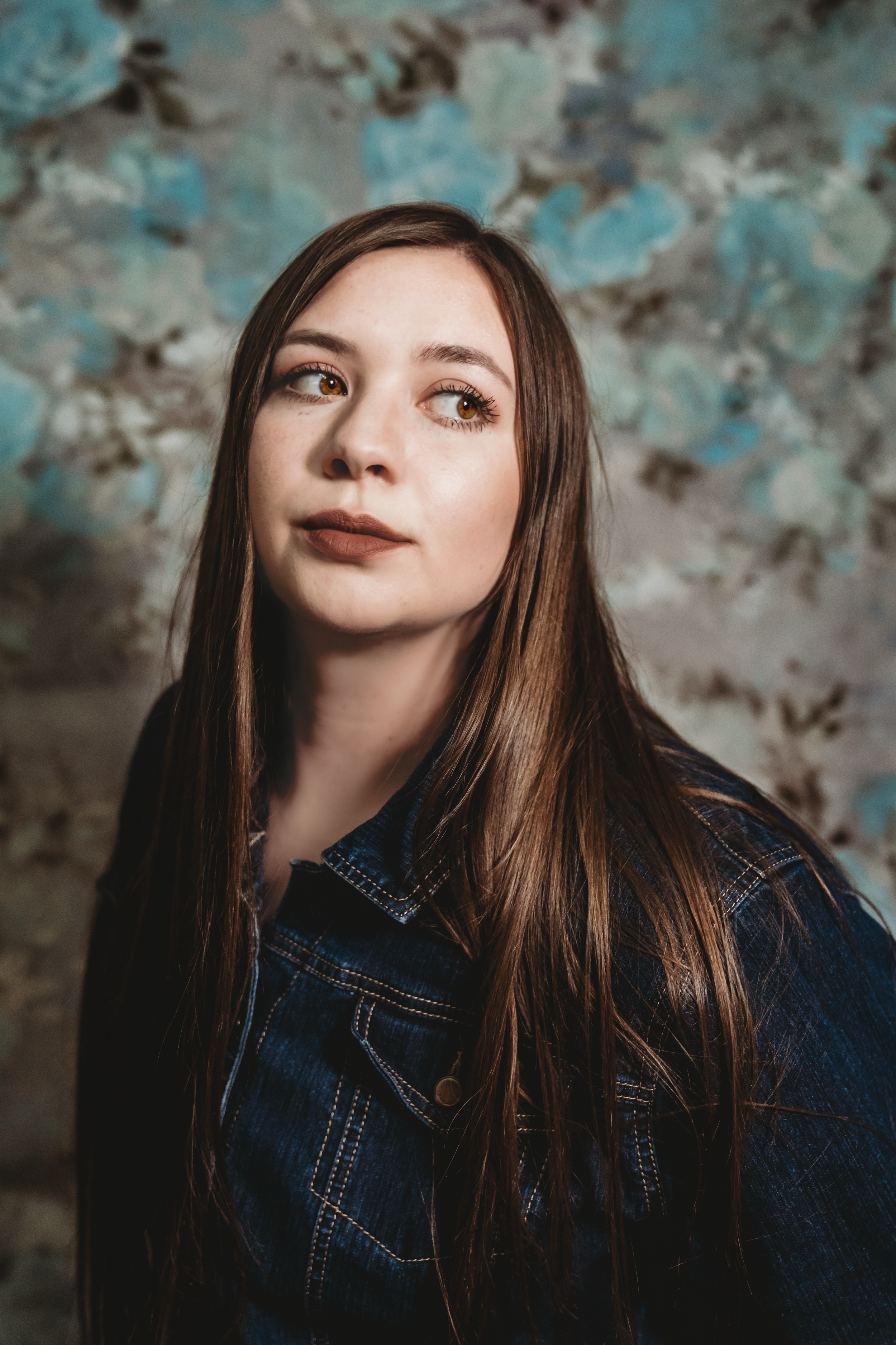 Young woman with long brown hair wearing a denim jacket, posing against a floral wallpaper background.