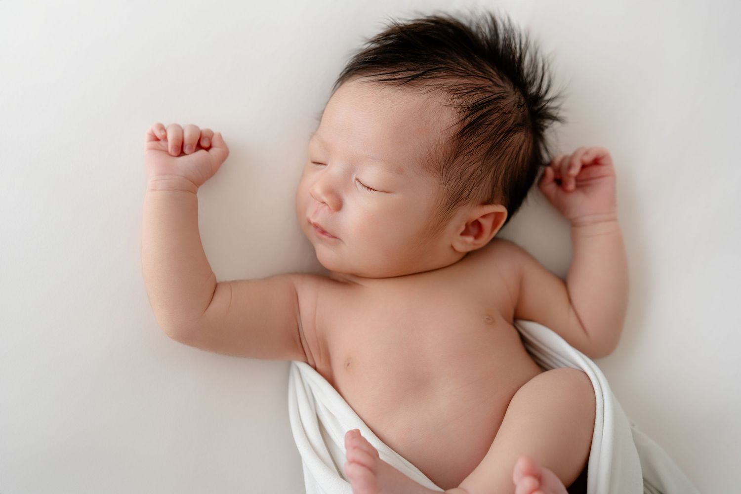 A sleeping newborn baby lies peacefully in soft white fabric creating a dreamy artistic composition.