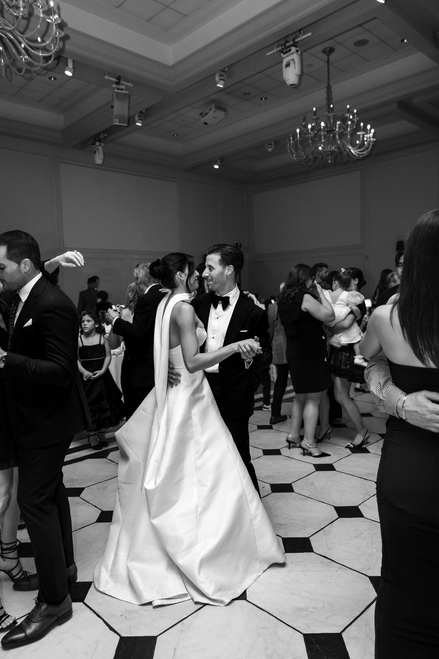 Black and white wedding reception photo showing couples dancing together in an elegant ballroom with chandeliers.