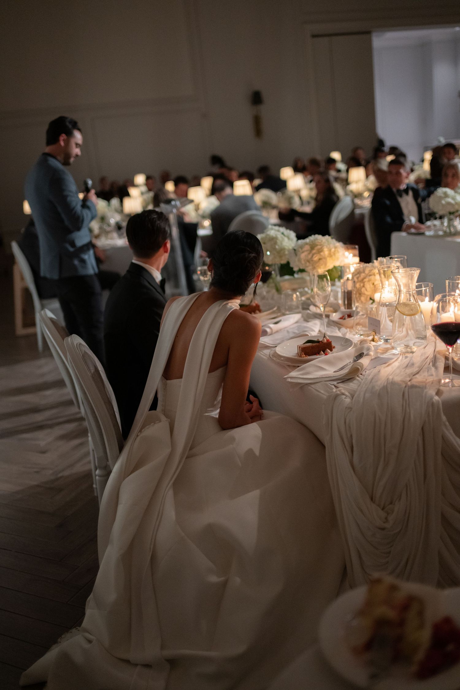 Elegant wedding reception table with white linens, glowing candles and floral centerpieces in a dimly lit ballroom.