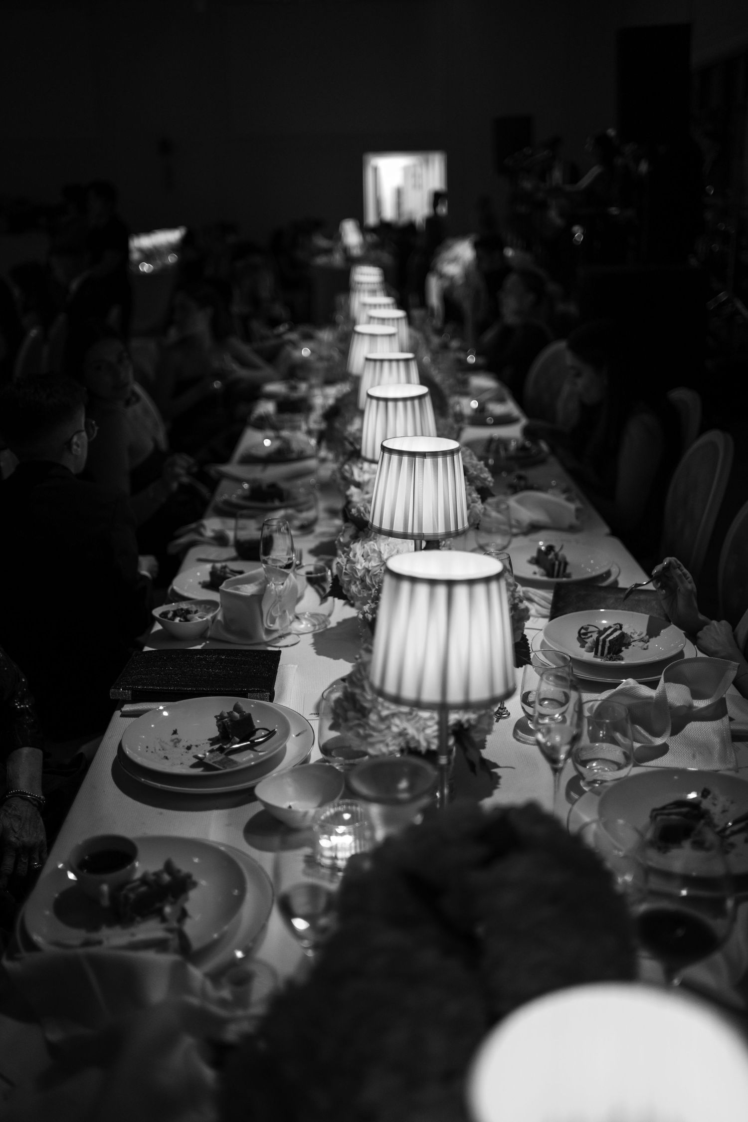 A series of elegant table settings at a formal dinner party illuminated by glowing white tiered lampshades.