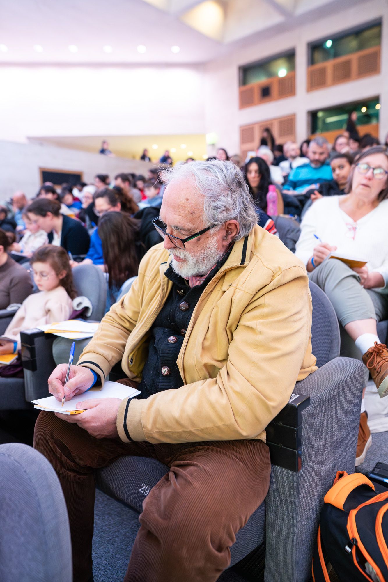 A person in a tan jacket takes notes while sitting in a crowded conference room.