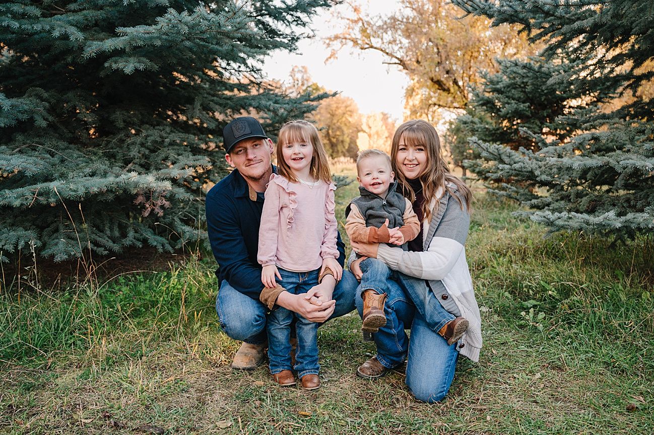 A family sits together on grass surrounded by evergreen trees during a golden sunset photo session.