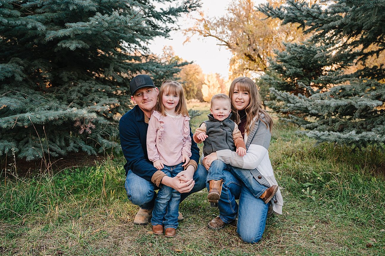 A family portrait taken between evergreen trees during golden hour with a warm sunset glow in the background.