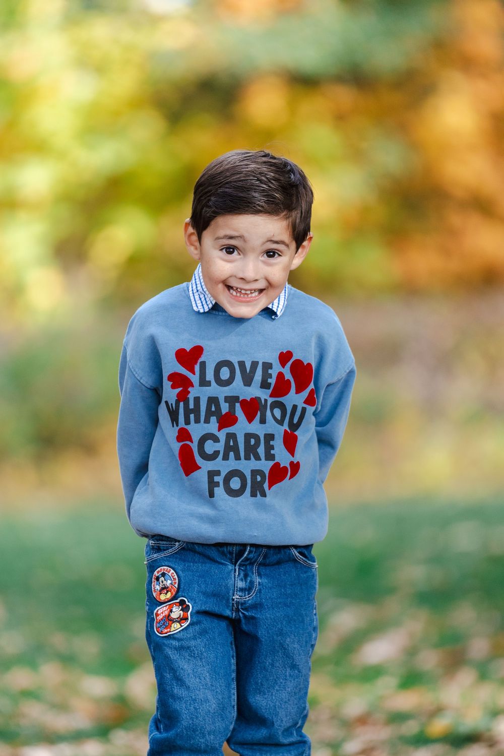 A child wearing a blue sweater with hearts and text stands in fallen autumn leaves outdoors.
