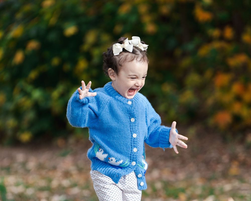 Child wearing blue cardigan and polka dot pants plays joyfully in autumn leaves on ground.