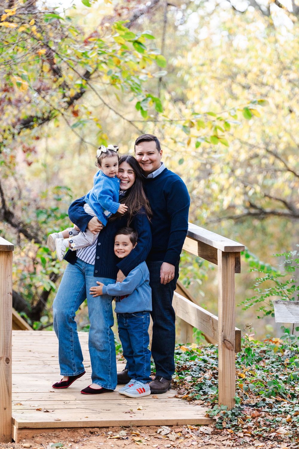 A family shares playful moments together on a wooden boardwalk surrounded by autumn foliage.