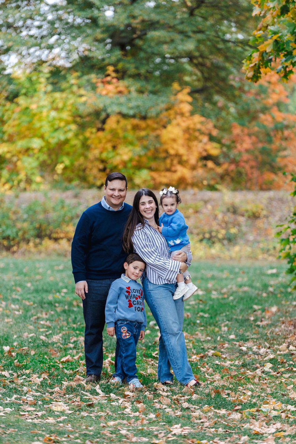 A family enjoying a fall day together on grass covered with colorful autumn leaves in a park setting.
