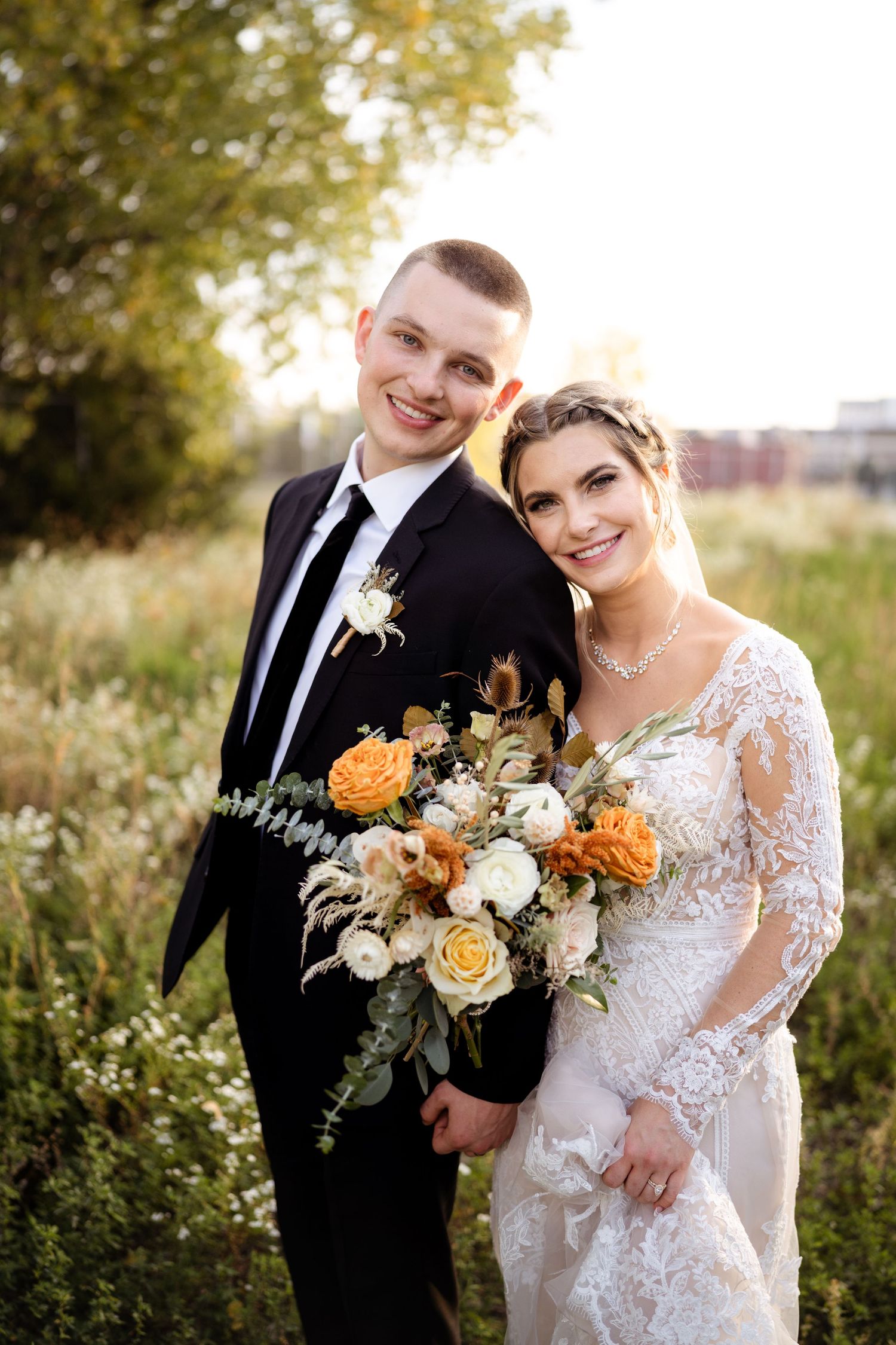 Smiling bride and groom pose together in a golden field holding an orange and white bouquet.