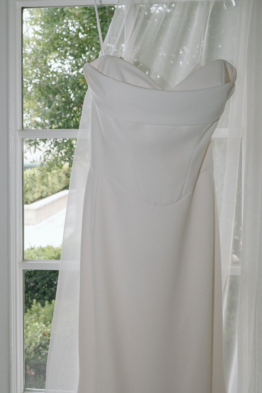 A white strapless wedding dress hangs elegantly by a window with sheer curtains and natural light.