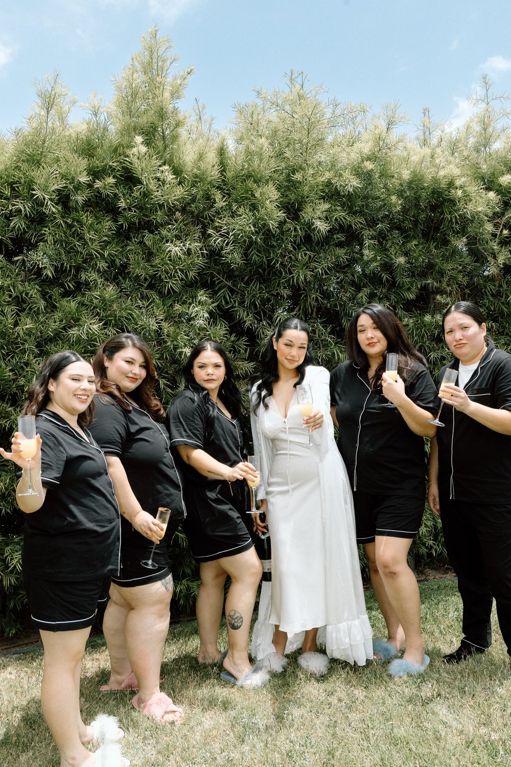Wedding party in black and white attire posing together on grass with hedge backdrop.