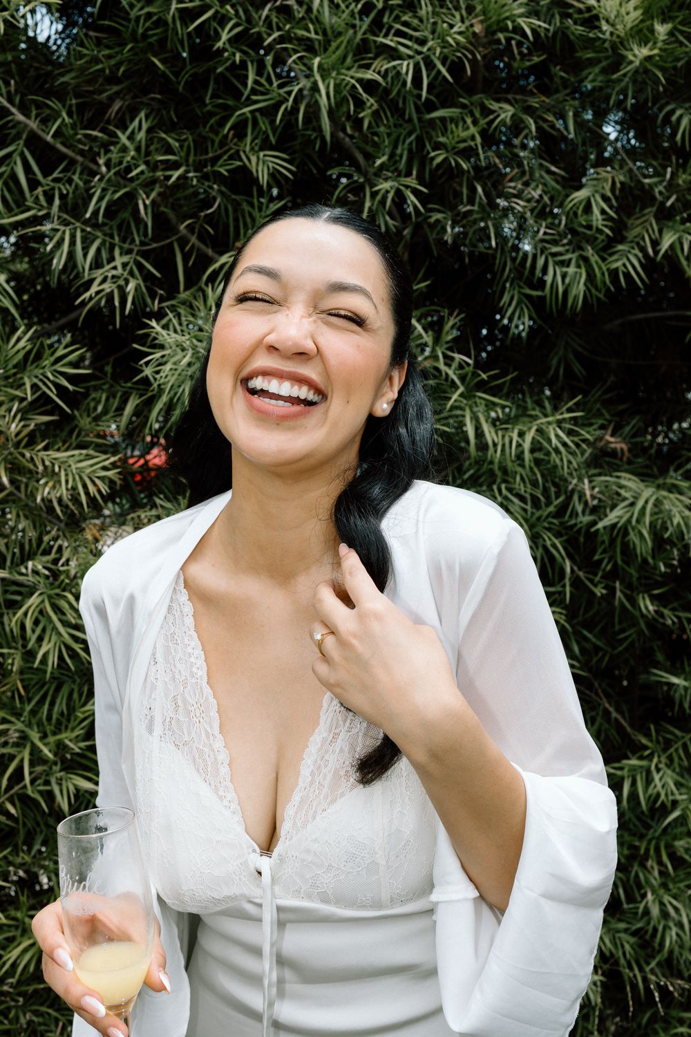 Person wearing white lace top smiling joyfully in front of green foliage.
