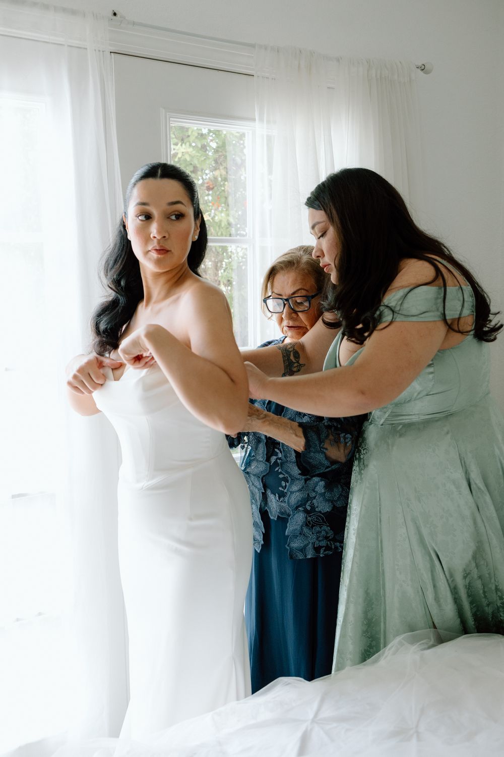 Wedding dress fitting scene in bright sunlit room with white curtains.