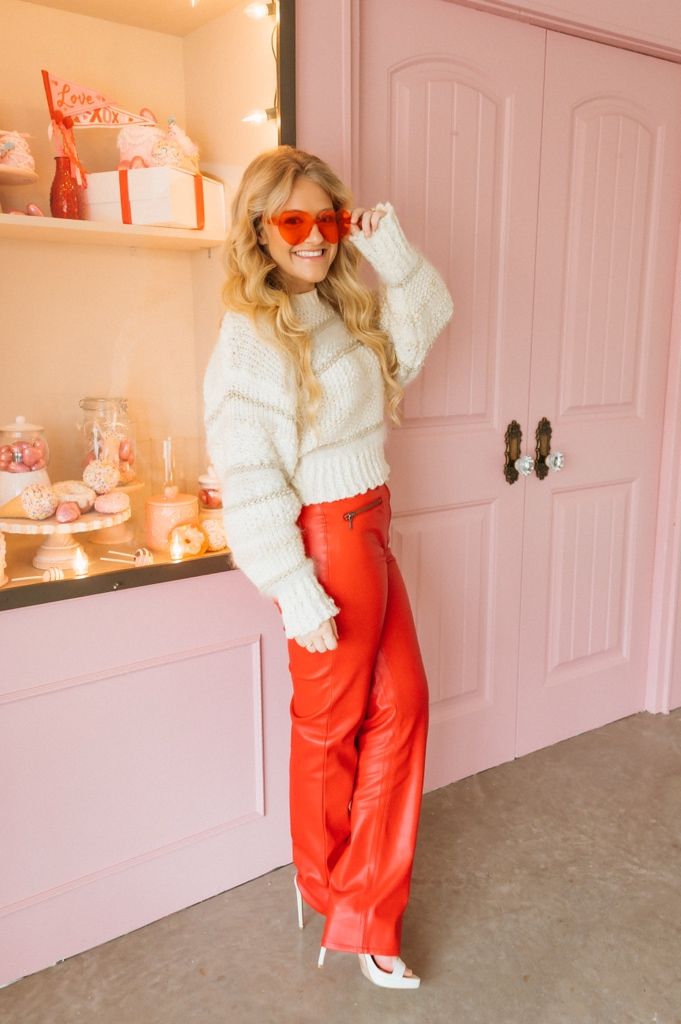 Person wearing white sweater and red pants posing against pink wall with decorative shelving.