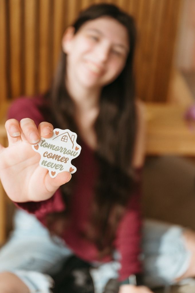Someone holding up a sticker with 'tomorrow comes never' text against a wooden slat background.
