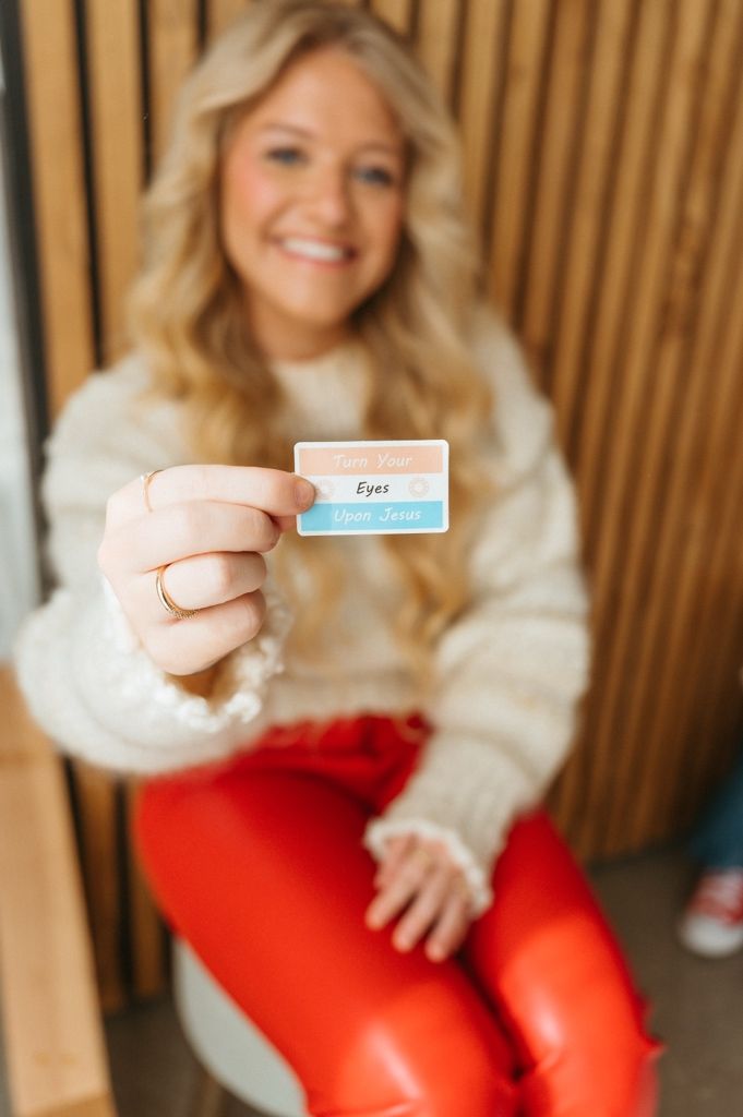 Someone in a white sweater and red pants holds up a small blue card against a wooden wall backdrop.