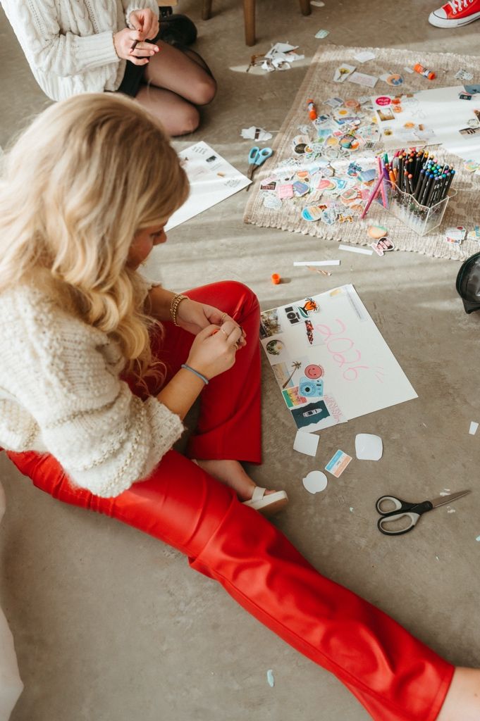 Person in white sweater and red pants working on craft project with scattered supplies on floor.
